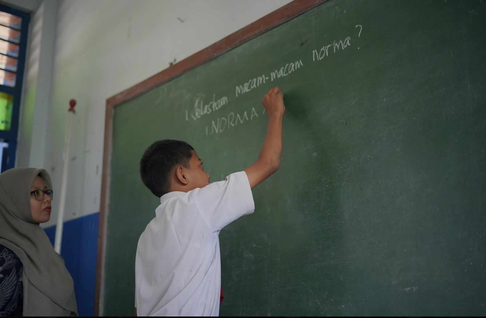 Siswa sekolah dasar berlatih menulis di papan tulis, didampingi gurunya